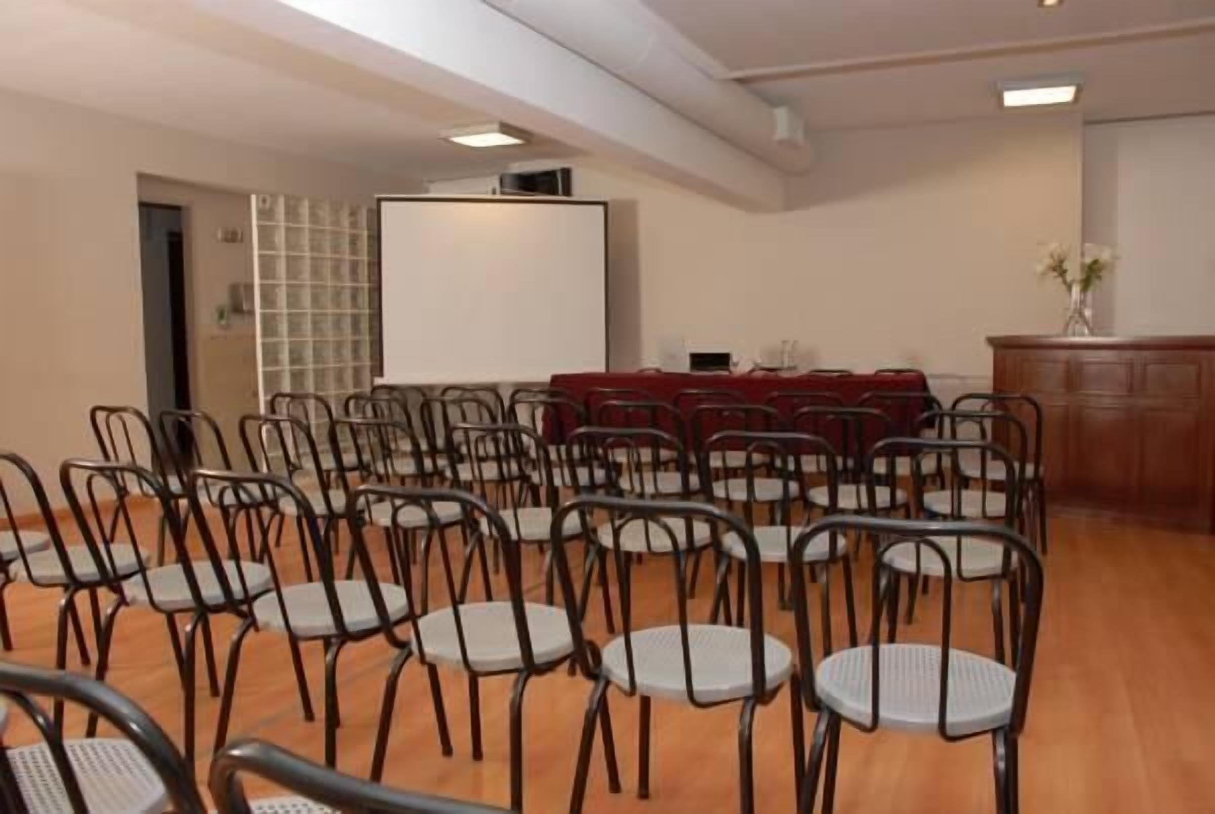 Un salón de conferencias con hileras de sillas, una pantalla de proyector y una mesa con mantel rojo.