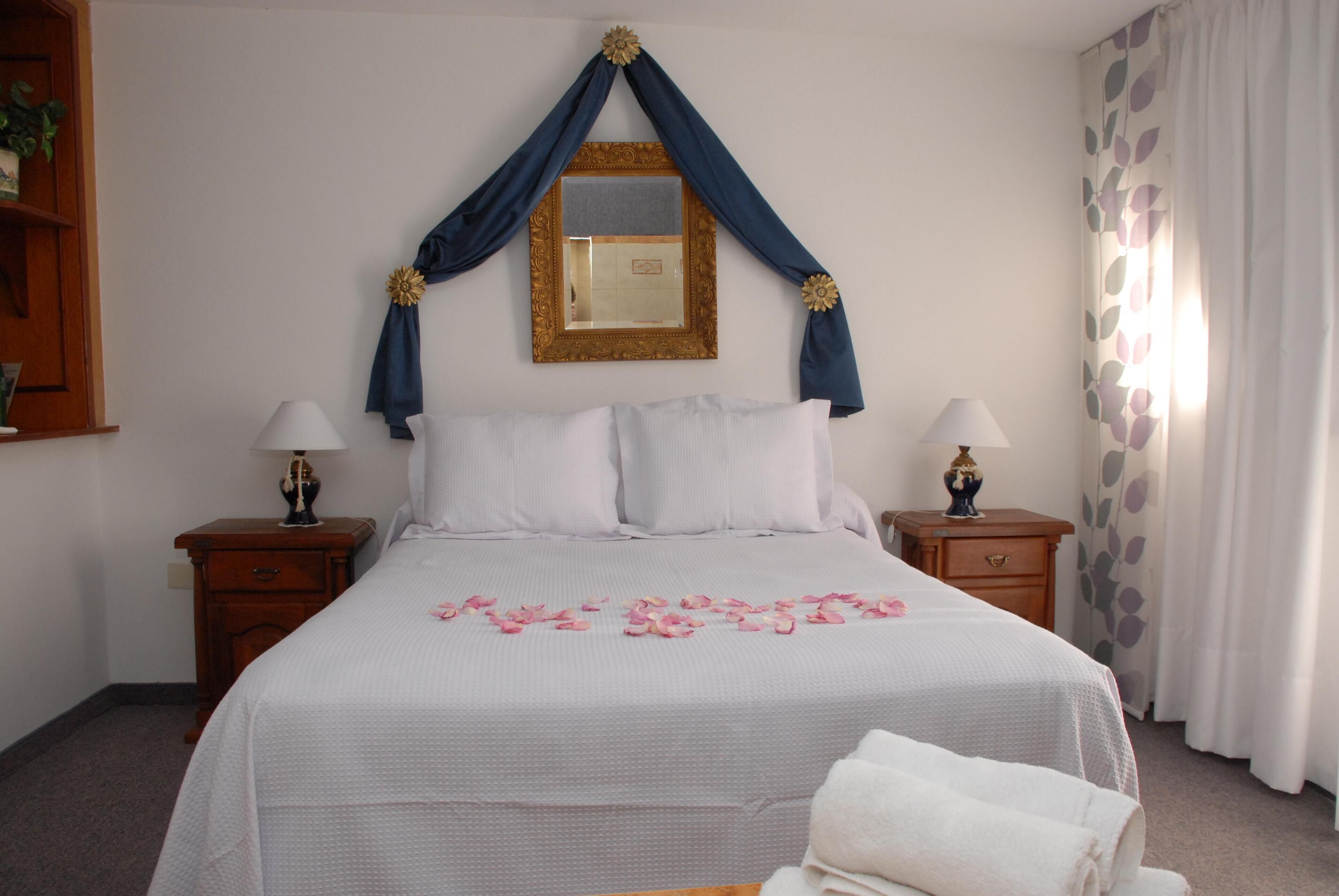 A neatly made bed with a white bedspread, two pillows, and a decorative mirror with draped curtains above. There are two bedside tables with lamps, a wooden nightstand, and a window with a curtain.