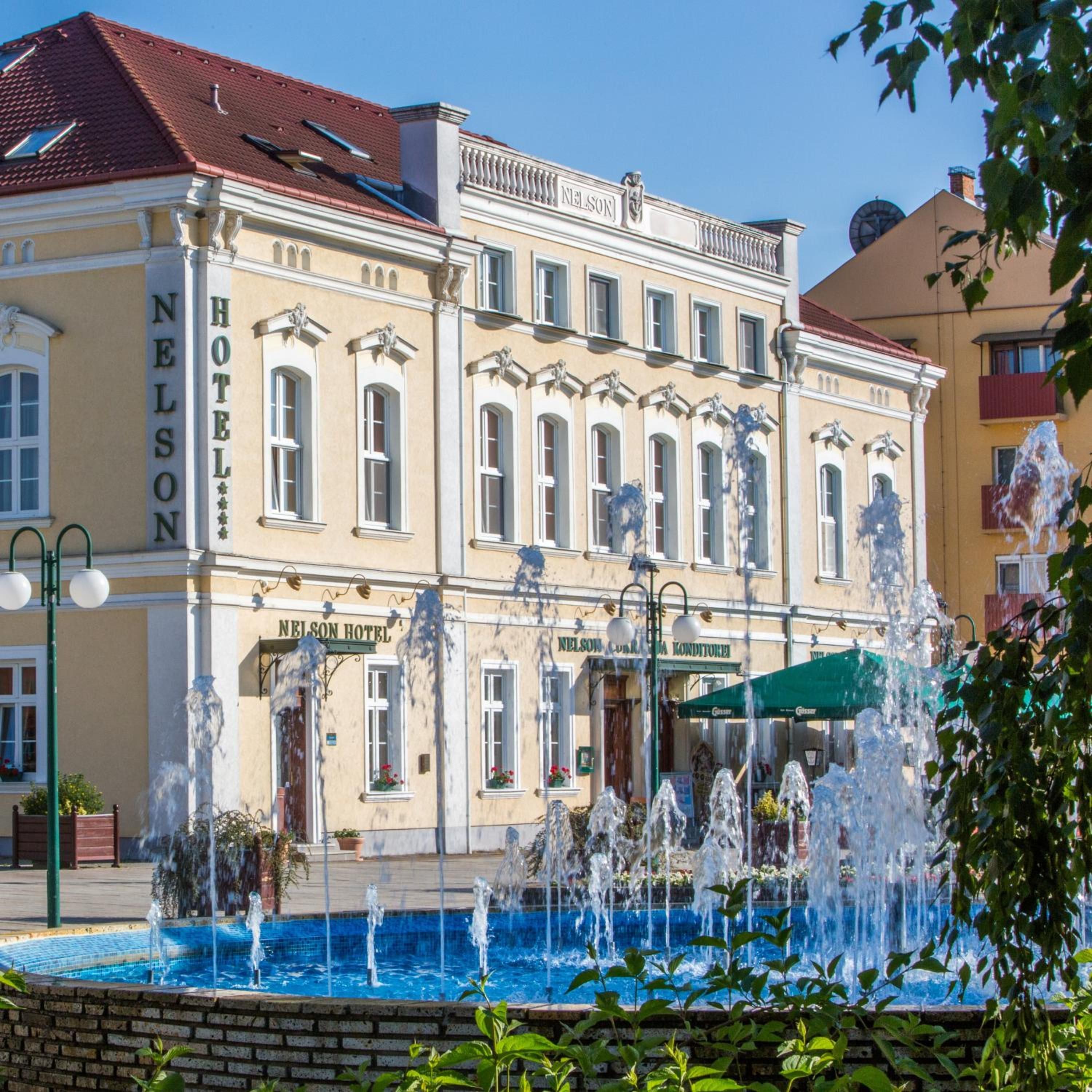 A hotel with a fountain in front, a red roof, and a sign that reads 'Nelson Hotel'.