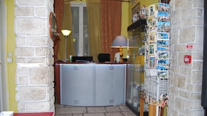 A room with a stone wall, a red table, a desk with a computer, and a wall-mounted rack of postcards. - Azur (Nice)