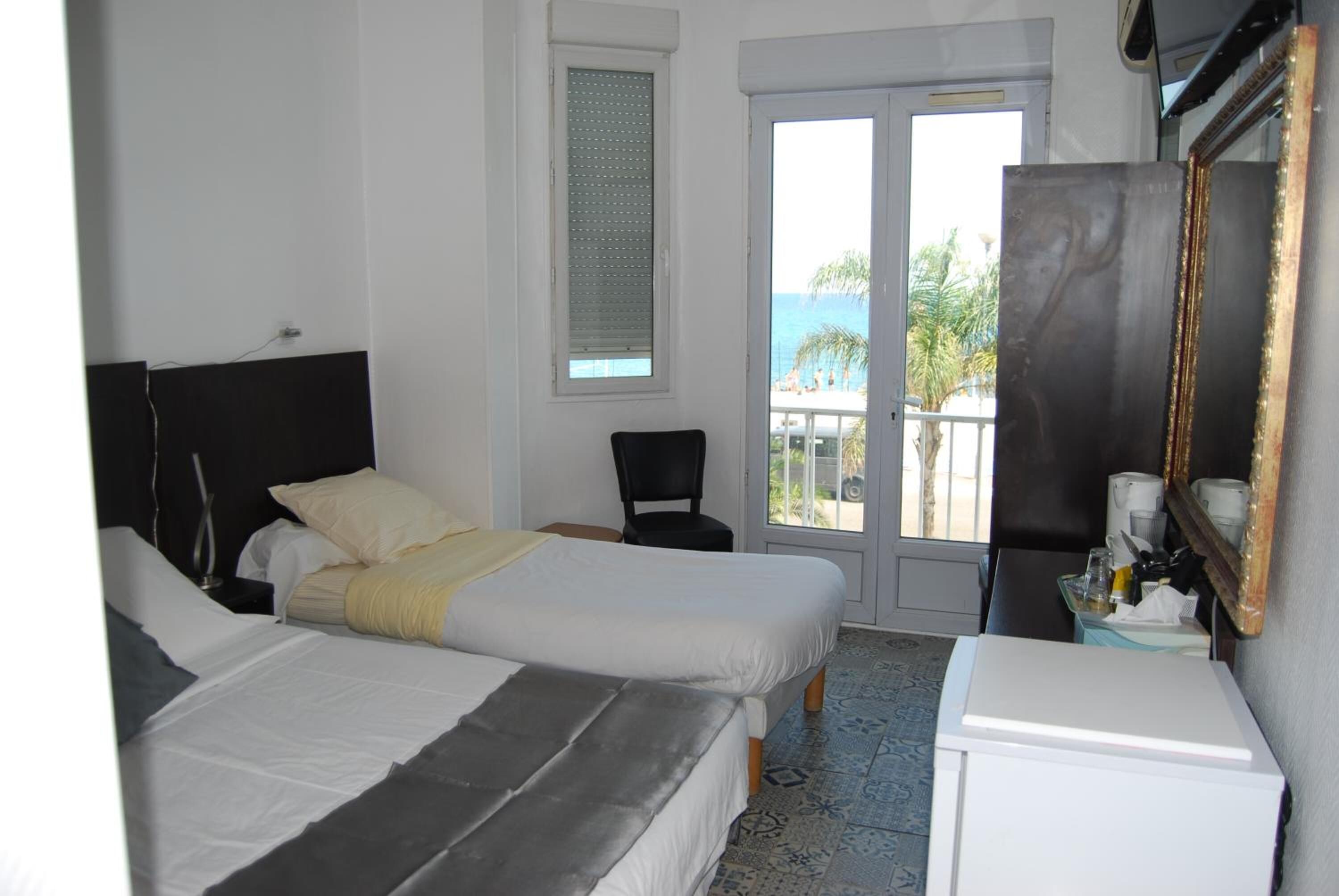 Une chambre d’hôtel avec deux lits, un balcon donnant sur la mer, une petite table avec une lampe et un miroir au mur.