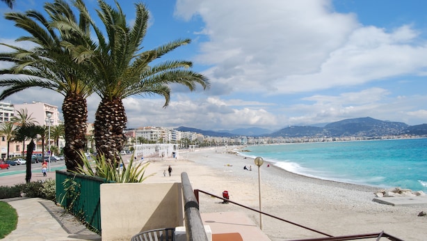 A beach with palm trees, a staircase leading down to the beach, and a view of the ocean and distant mountains. - Azur (Nice)