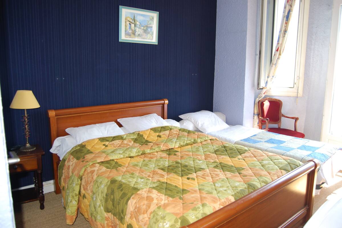Una cama doble con colcha floral, cabecera de madera, mesita de noche con lámpara y un cuadro en la pared.