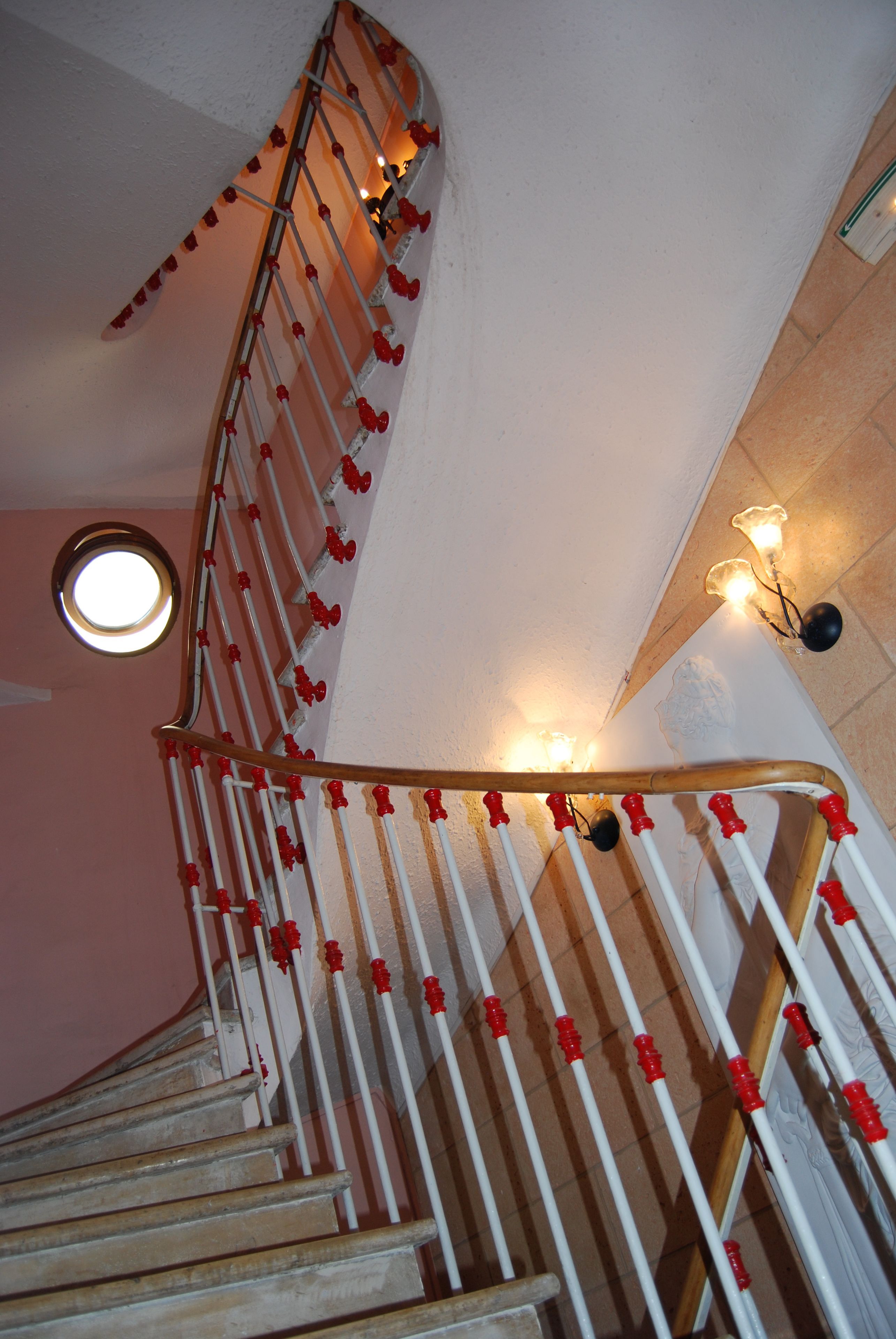 A spiral staircase with red and white balusters, a round window at the top, and wall-mounted lights.