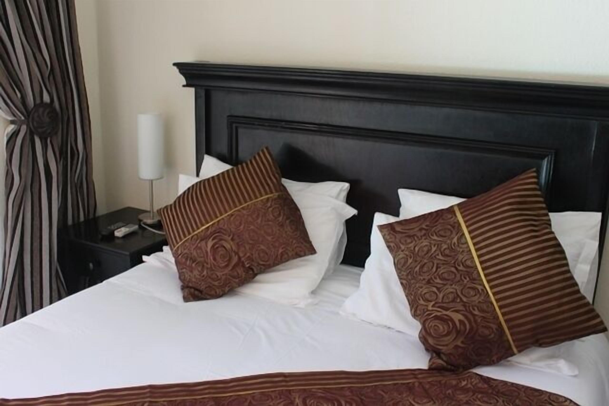 Een net opgemaakt bed met witte lakens en gestreepte kussens, een donker hoofdbord en een nachtkastje met een lamp en een afstandsbediening.