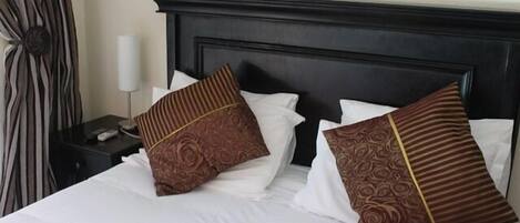Una cama bien hecha con sábanas blancas y almohadas estampadas, un cabecero oscuro y una mesita de noche con una lámpara y un control remoto.