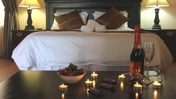 Una cama bien hecha, con almohadas y una botella de vino sobre una mesita de noche, acompañada de velas y un cuenco de fruta.
