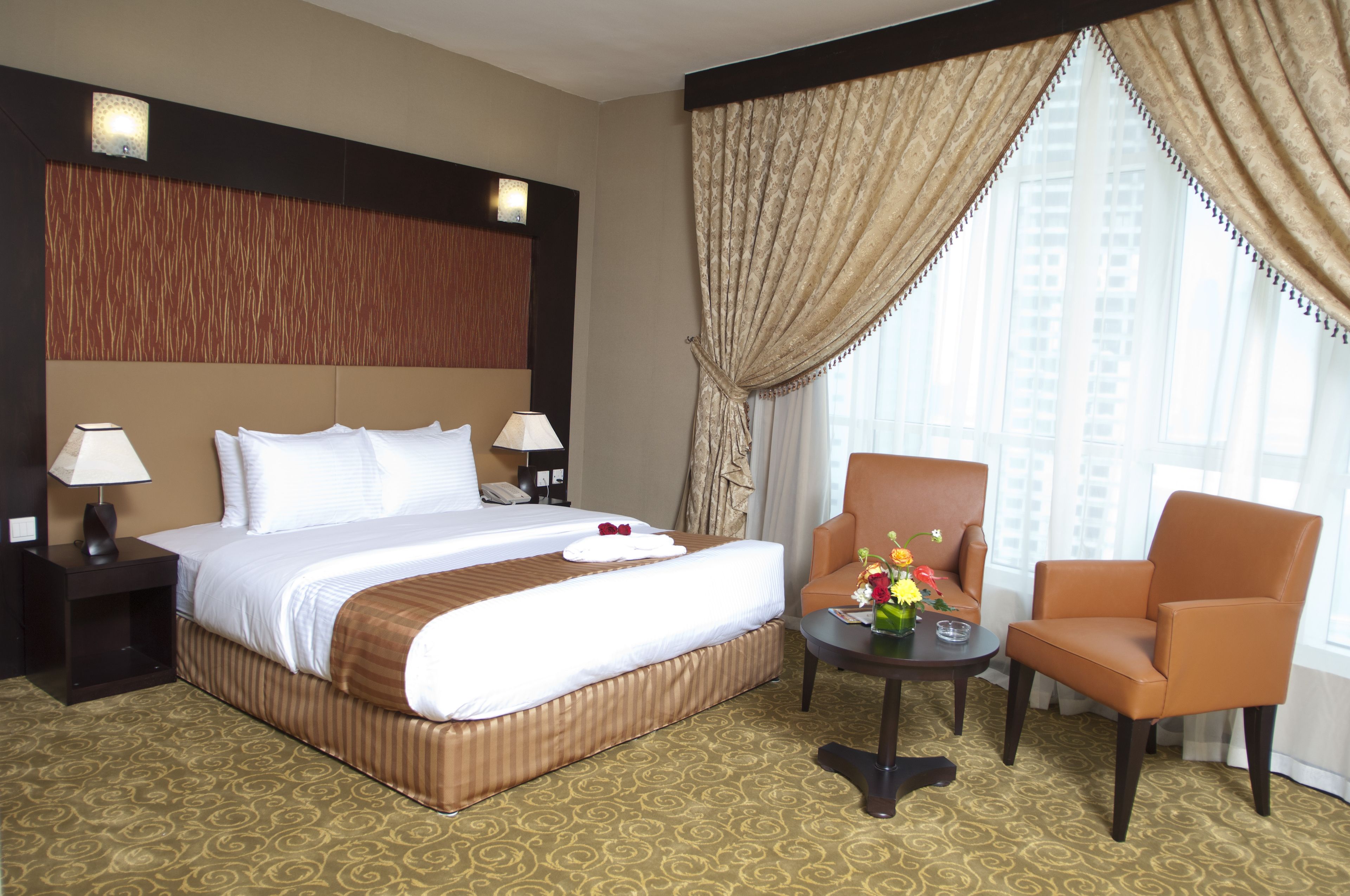 Une chambre d’hôtel avec un grand lit, deux tables de chevet avec des lampes, une petite table sur laquelle est posé un vase de fleurs, et une fenêtre avec des rideaux.