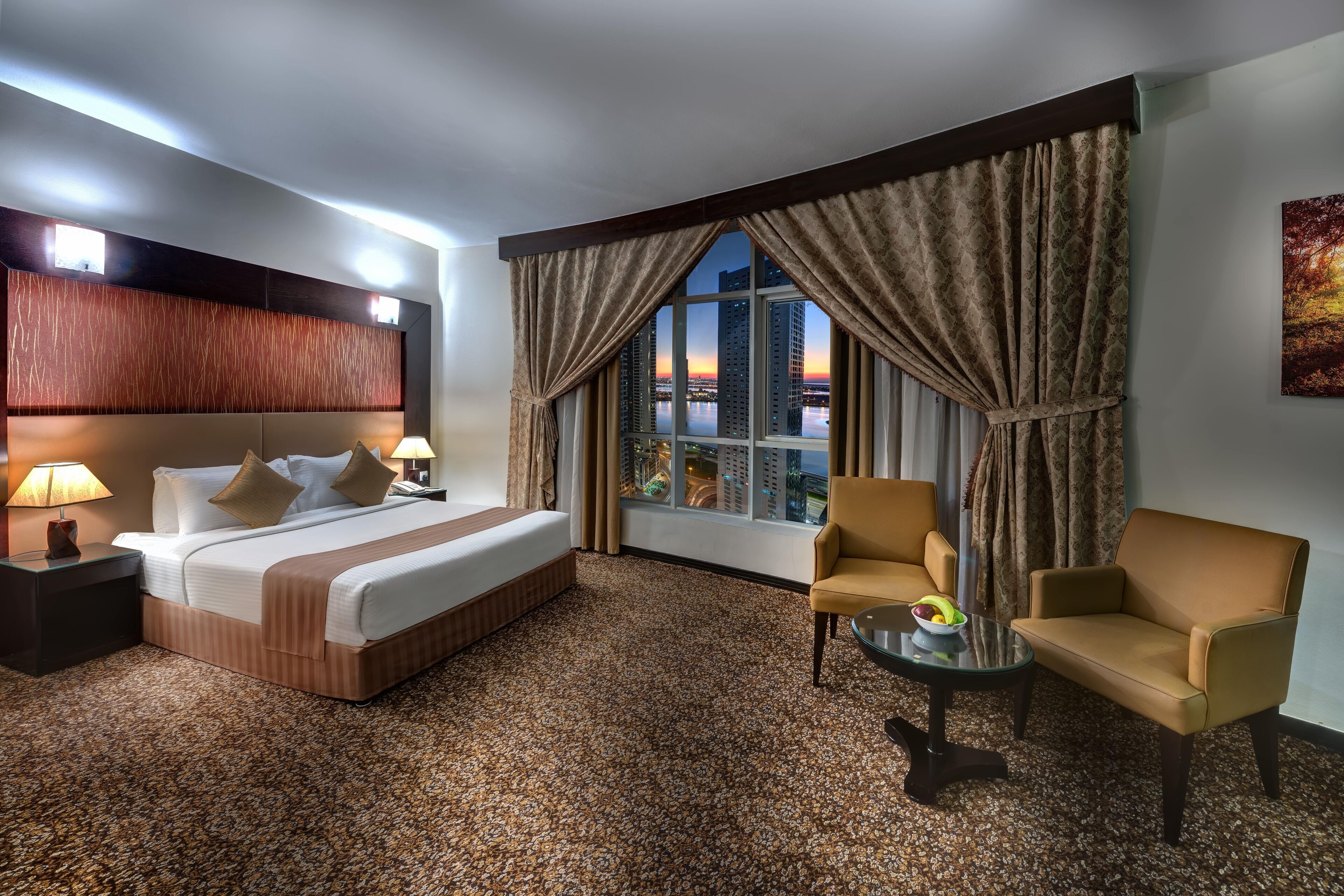 Quarto de hotel com uma cama grande, duas cadeiras, uma mesinha e vista para a cidade ao pôr do sol.