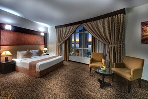 A hotel room with a large bed, two chairs, a small table, and a view of the cityscape at sunset. - Aryana Hotel (Sharjah)