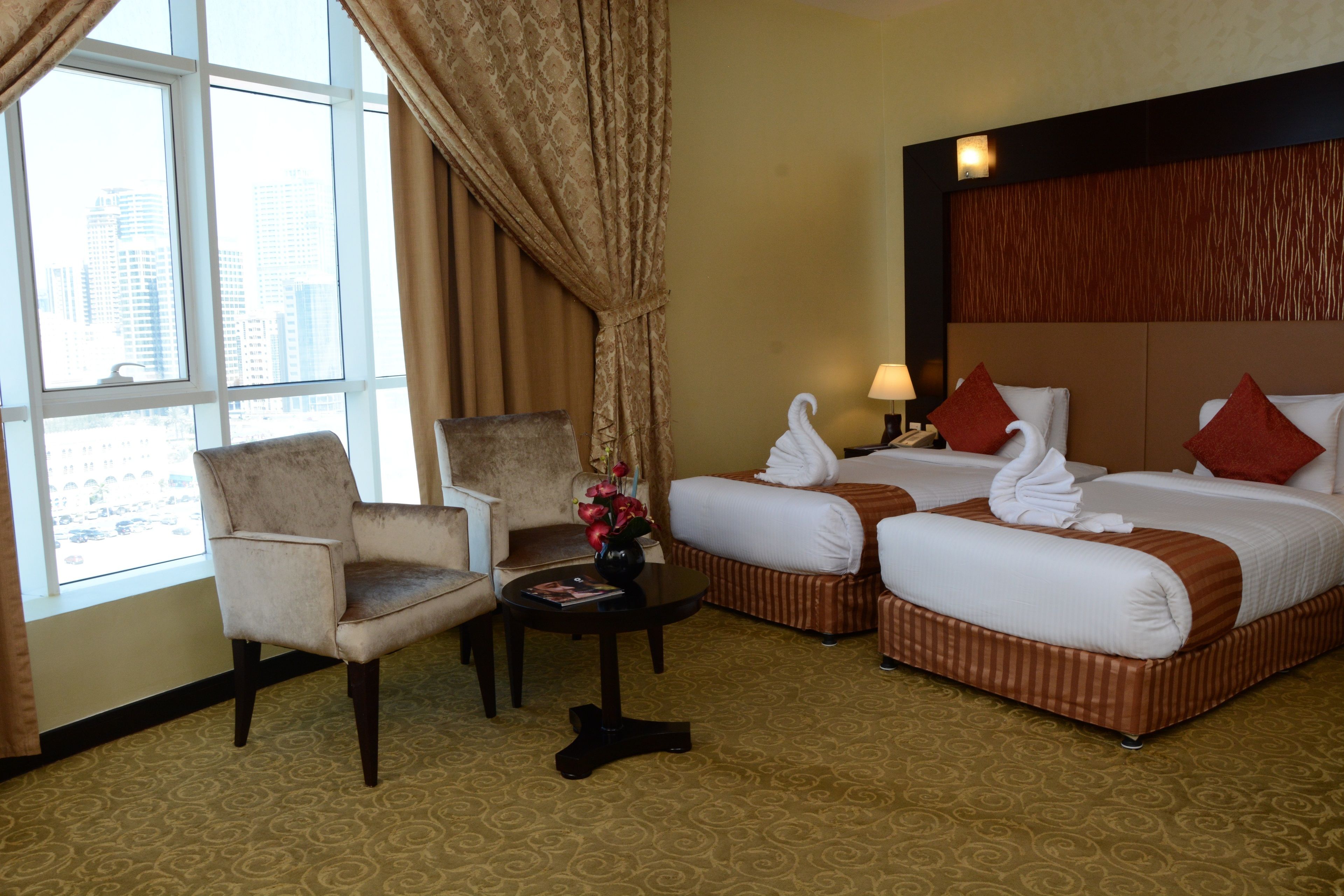 Ein Hotelzimmer mit zwei Betten, einem Sessel, einem kleinen Tisch mit einer Vase und Blick auf die Stadt durch große Fenster.