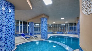 Ein Hallenbad mit blauen Mosaikfliesen, Liegestühlen und großen Fenstern.