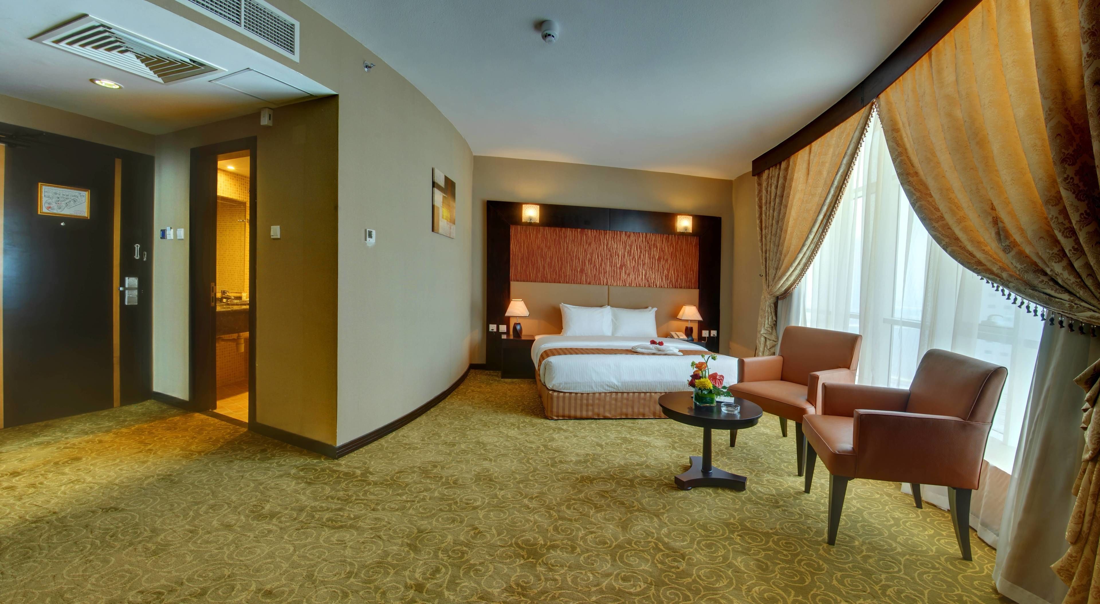 A hotel room with a bed, two bedside tables with lamps, a small round table with a vase, a chair, and a large window with curtains.
