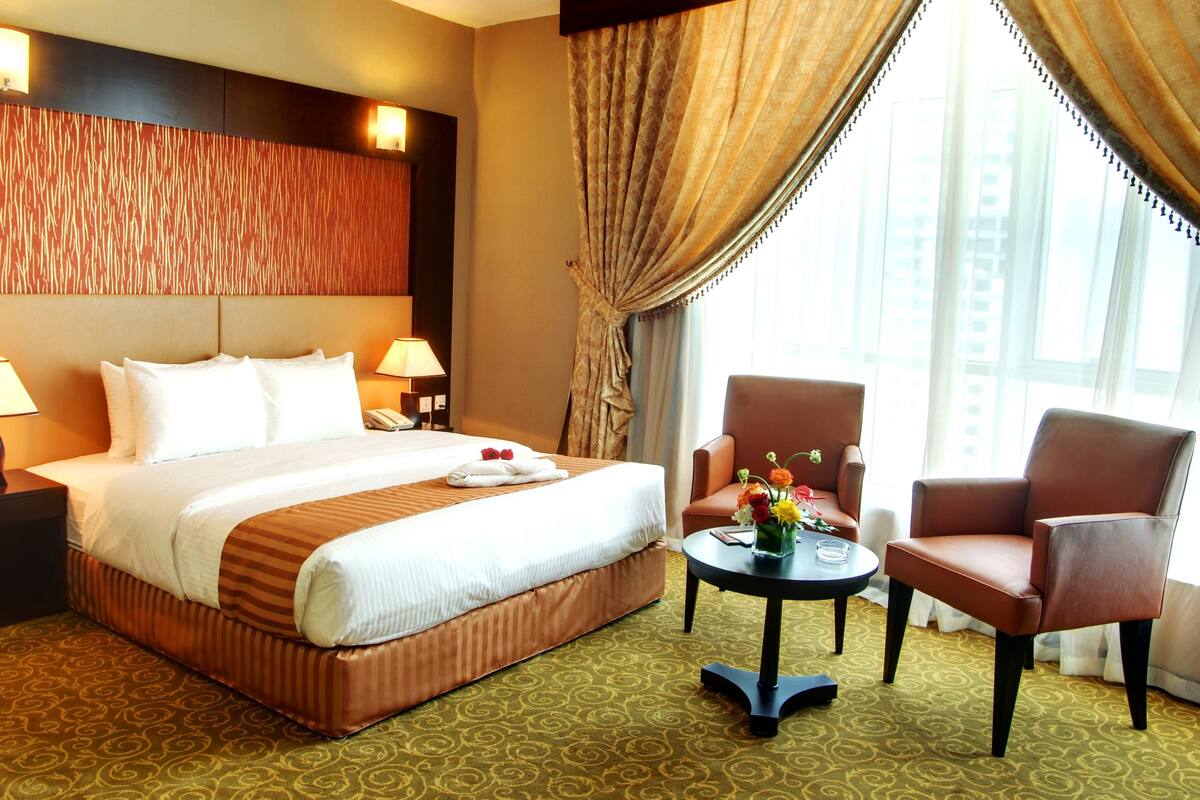 A hotel room with a large bed, two bedside lamps, a small table with a vase of flowers, two chairs, and a view of the city through large windows.