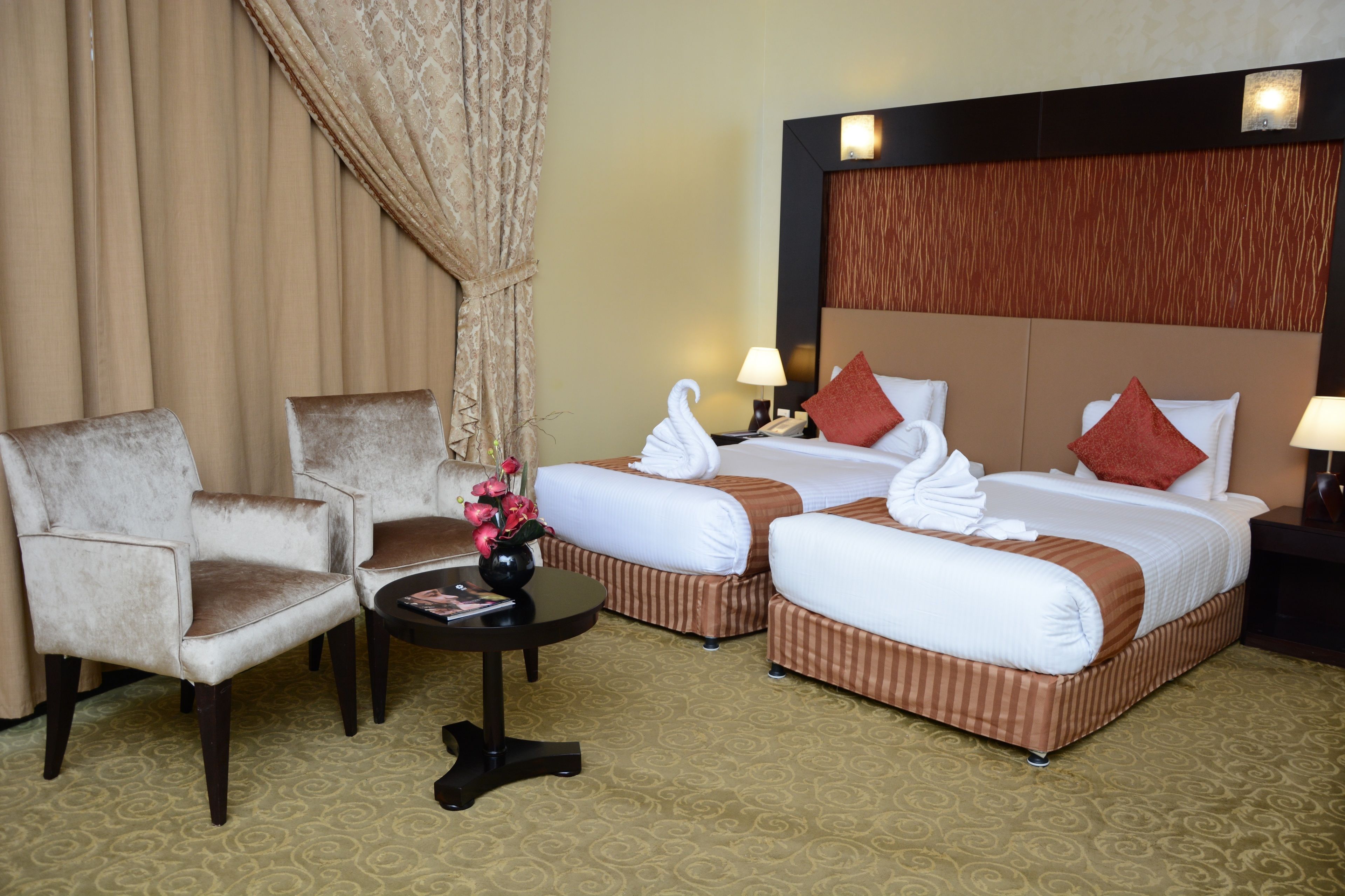 A hotel room with two beds, a sitting area with chairs, a small table with a vase of flowers, and a nightstand with a lamp.