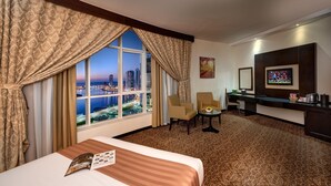 Ein Hotelzimmer mit großem Fenster und Blick auf die Stadt, einem Bett mit einer Zeitschrift, einem kleinen Tisch mit einer Schale, zwei Sesseln, einem Schreibtisch mit Fernseher und einer Kaffeemaschine.
