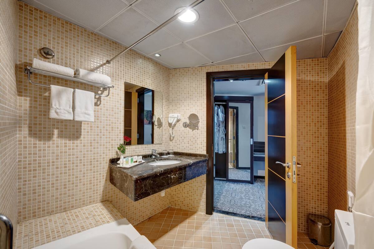 A bathroom with a tiled wall, a sink, a mirror, a towel rack, and a door leading to another room.