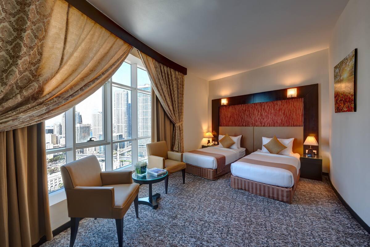 A hotel room with two beds, a sitting area with a table, and a large window offering a city view.