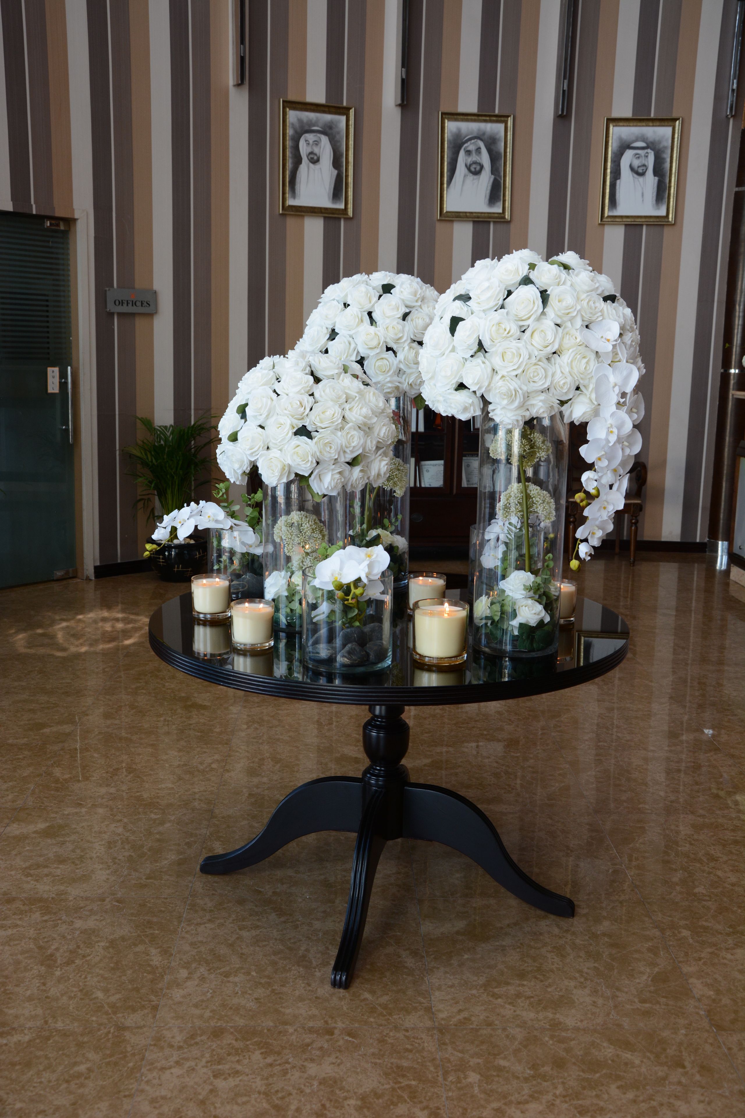 Uma mesa com vasos de flores brancas e velas, em frente a uma parede listrada com quadros.