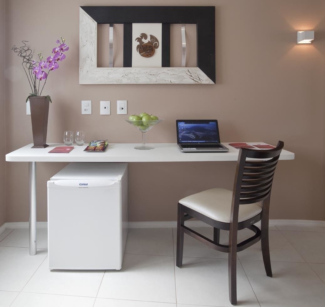 A compact kitchenette with a refrigerator, a laptop on a white countertop, and a chair.