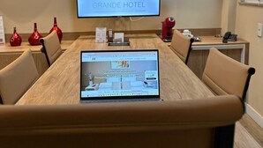 Une salle de conférence équipée d’une grande table, de chaises, d’un ordinateur portable et d’un téléviseur affichant le logo du Grand Hôtel. Des drapeaux sont accrochés au mur, au-dessus du téléviseur.