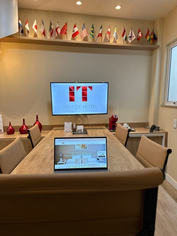 Una sala de conferencias con una mesa grande, sillas, una computadora portátil y un televisor que muestra el logotipo del Grande Hotel. Hay banderas colgadas en la pared sobre el televisor.