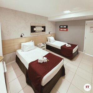 Habitación de hotel con dos camas, cada una con colchón blanco y cabecero oscuro. Hay una mesita de noche con lámpara, una obra de arte en la pared y una puerta con un cartel.