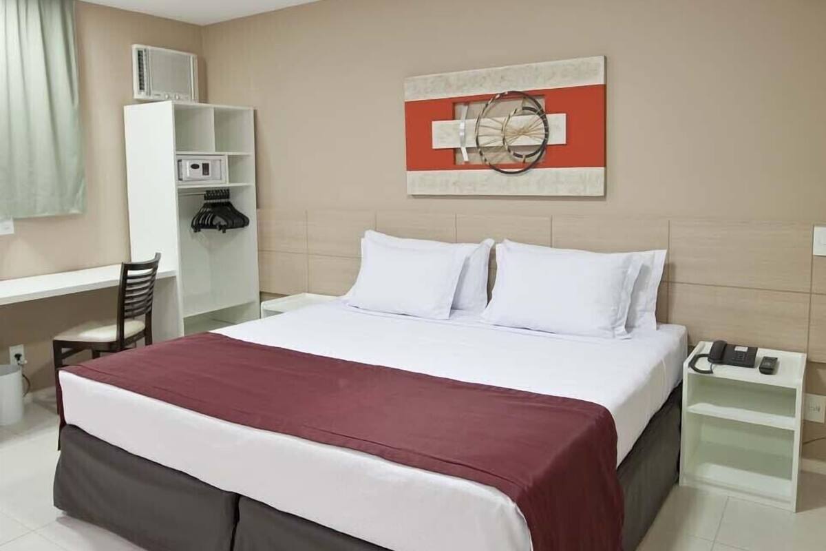Habitación de hotel con cama, escritorio, silla, armario y mesita de noche con teléfono.