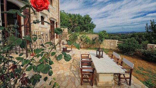 A stone building with a large red rose, outdoor seating, and a view of the landscape. - Strofilia Villas (Apokoronas)