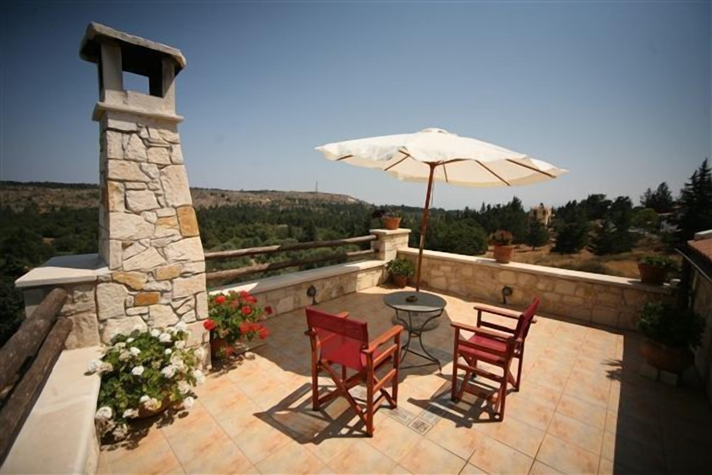 A patio with stone walls, a stone chimney, two wooden chairs, a small round table, and a white umbrella.