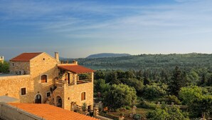 A stone building with a red-tiled roof, a balcony with potted plants, and a view of a forested hillside. - Strofilia Villas (Apokoronas)
