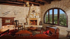 A rustic room with a stone fireplace, wooden furniture, and a view of the countryside through a large arched window. - Strofilia Villas (Apokoronas)