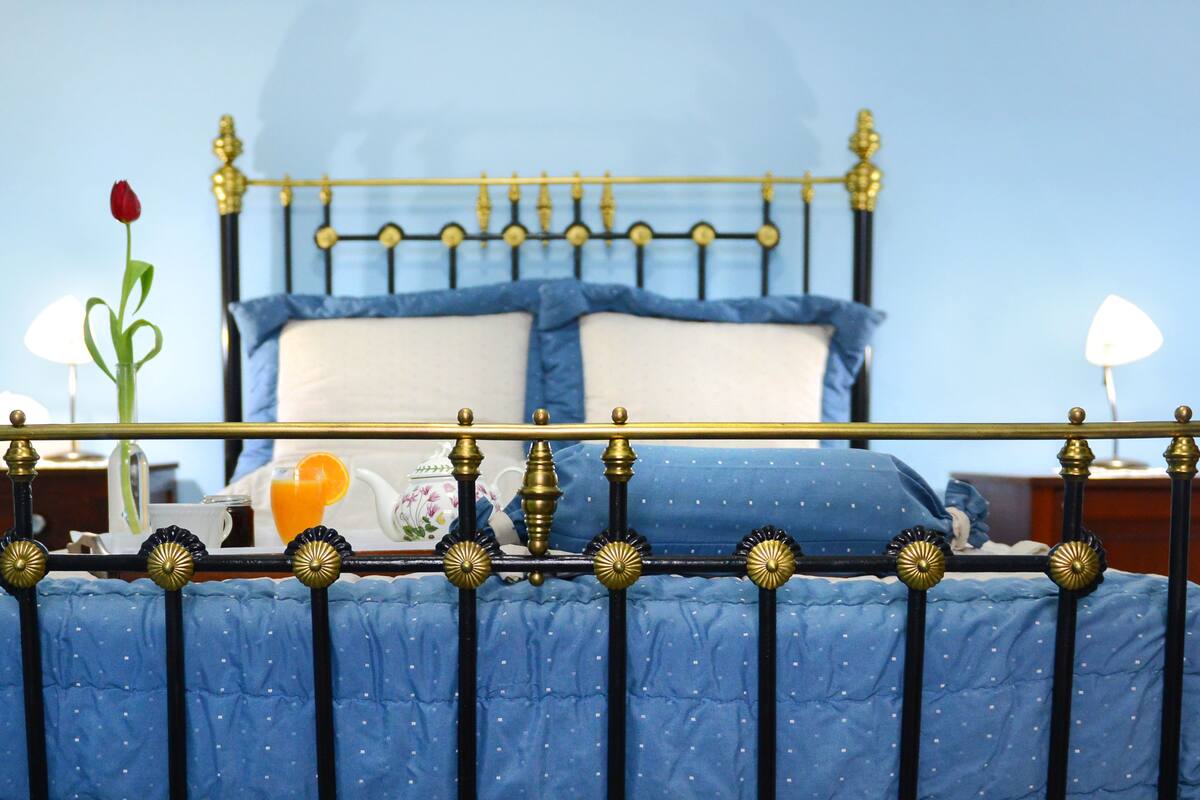 A bed with a blue quilt, a rose, a glass of orange juice, and a teapot on the nightstand.