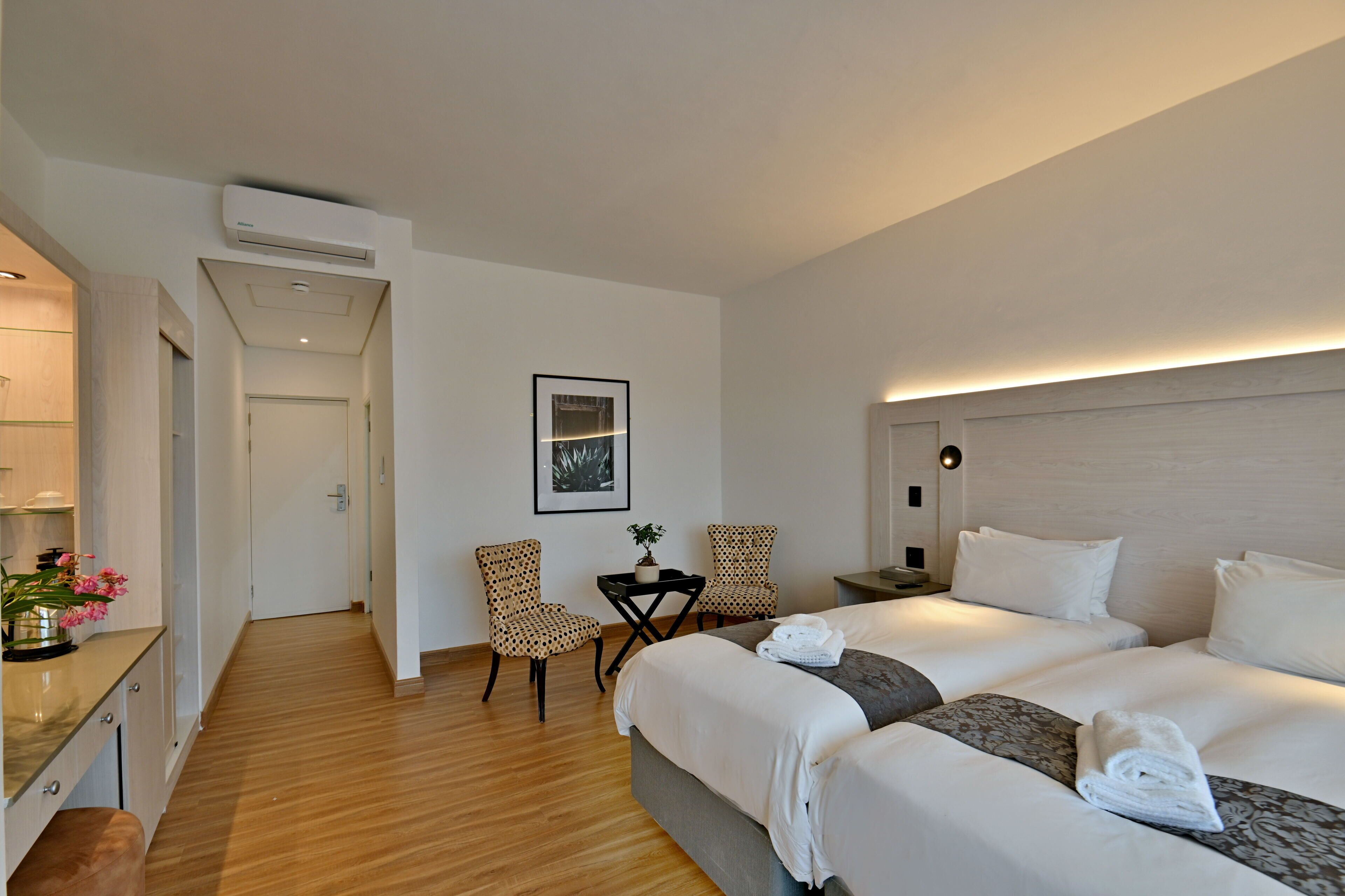 Habitación de hotel con piso de madera, dos camas, un escritorio con silla y una pequeña mesa con sillas.