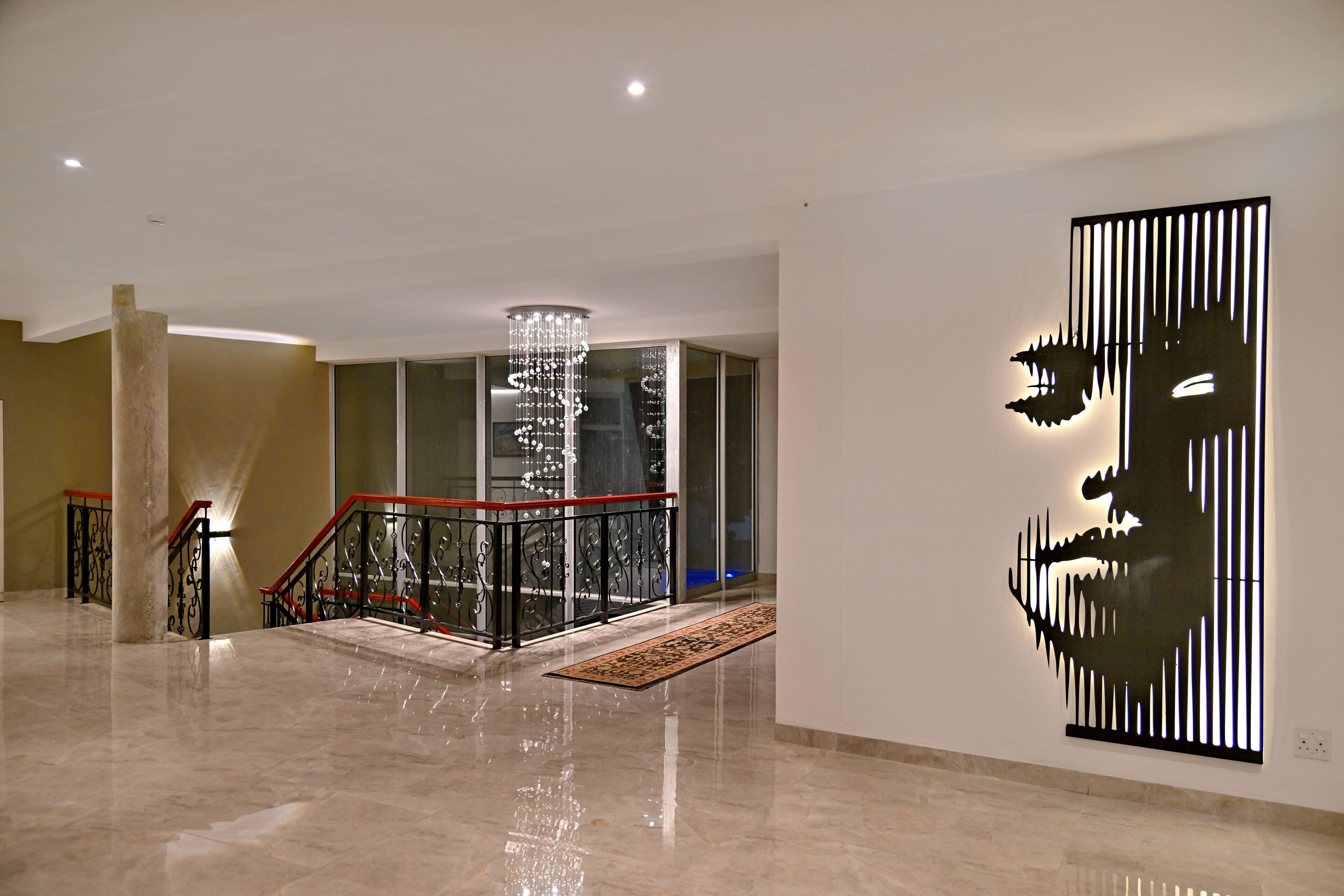 A spacious lobby with a polished marble floor, a grand staircase with a red railing, a modern chandelier, and a large abstract wall art piece.