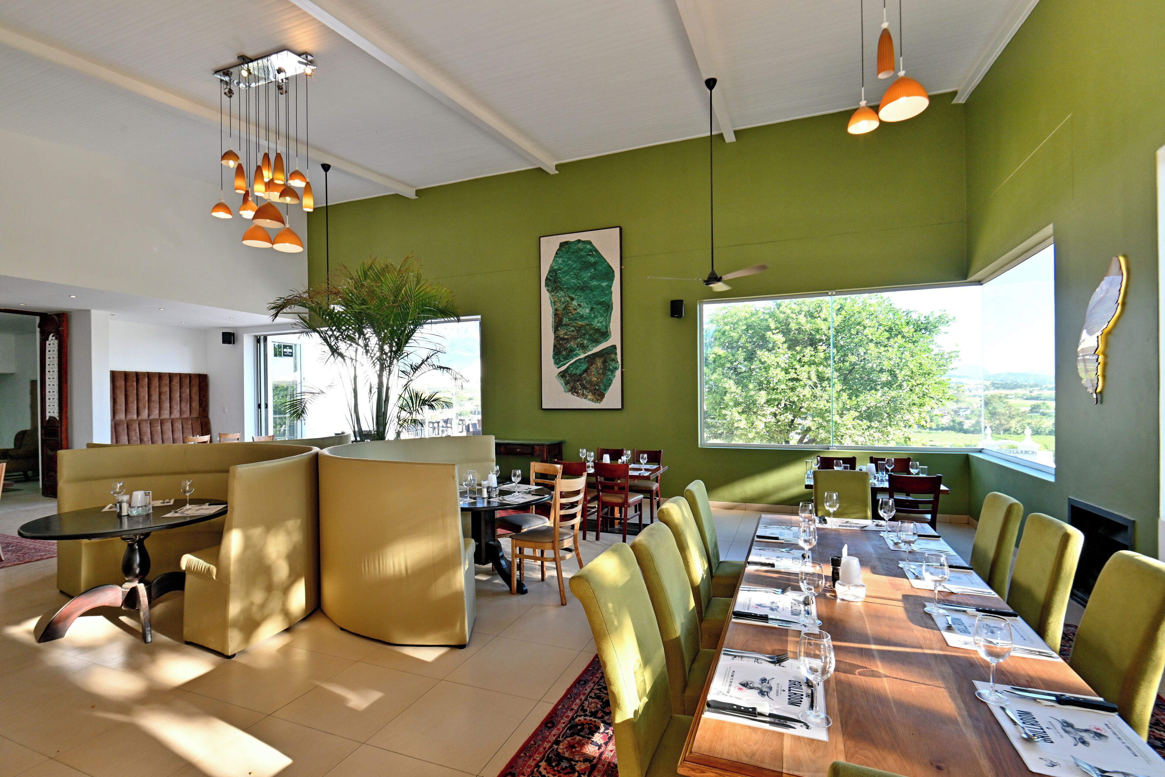 A modern dining area with a long wooden table, yellow chairs, and a large window offering a view of greenery.