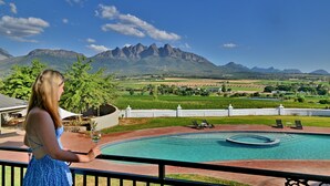 A pool area with a view of mountains, a person holding a glass, and lounge chairs. - Val Du Charron Wine & Leisure Estate (Wellington)