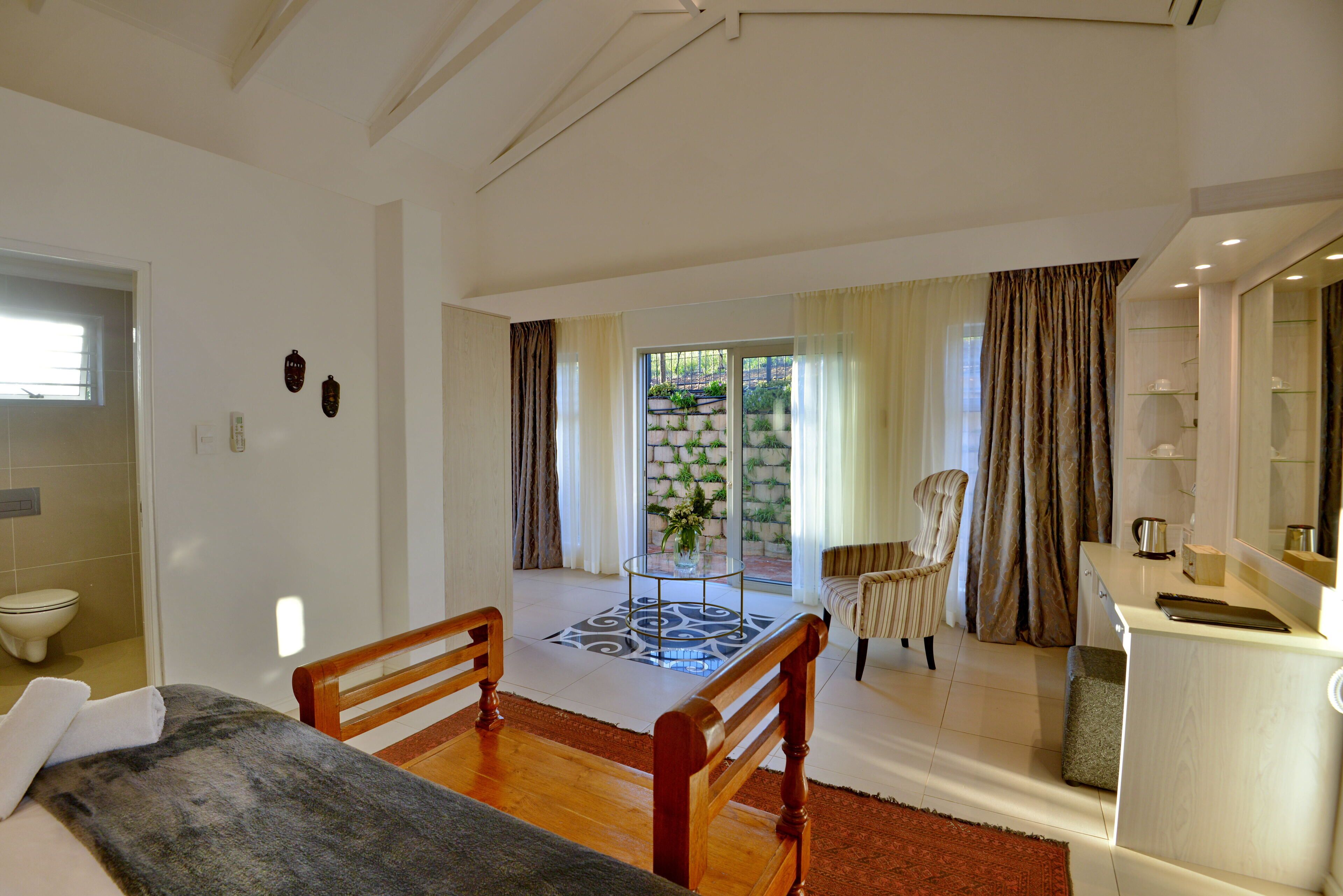 Habitación de hotel con cama, una silla de madera, una mesita con un jarrón, zona de cocina y vista a un jardín a través de un ventanal.