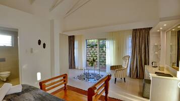 Habitación de hotel con cama, una silla de madera, una mesita con un jarrón, zona de cocina y vista a un jardín a través de un ventanal.