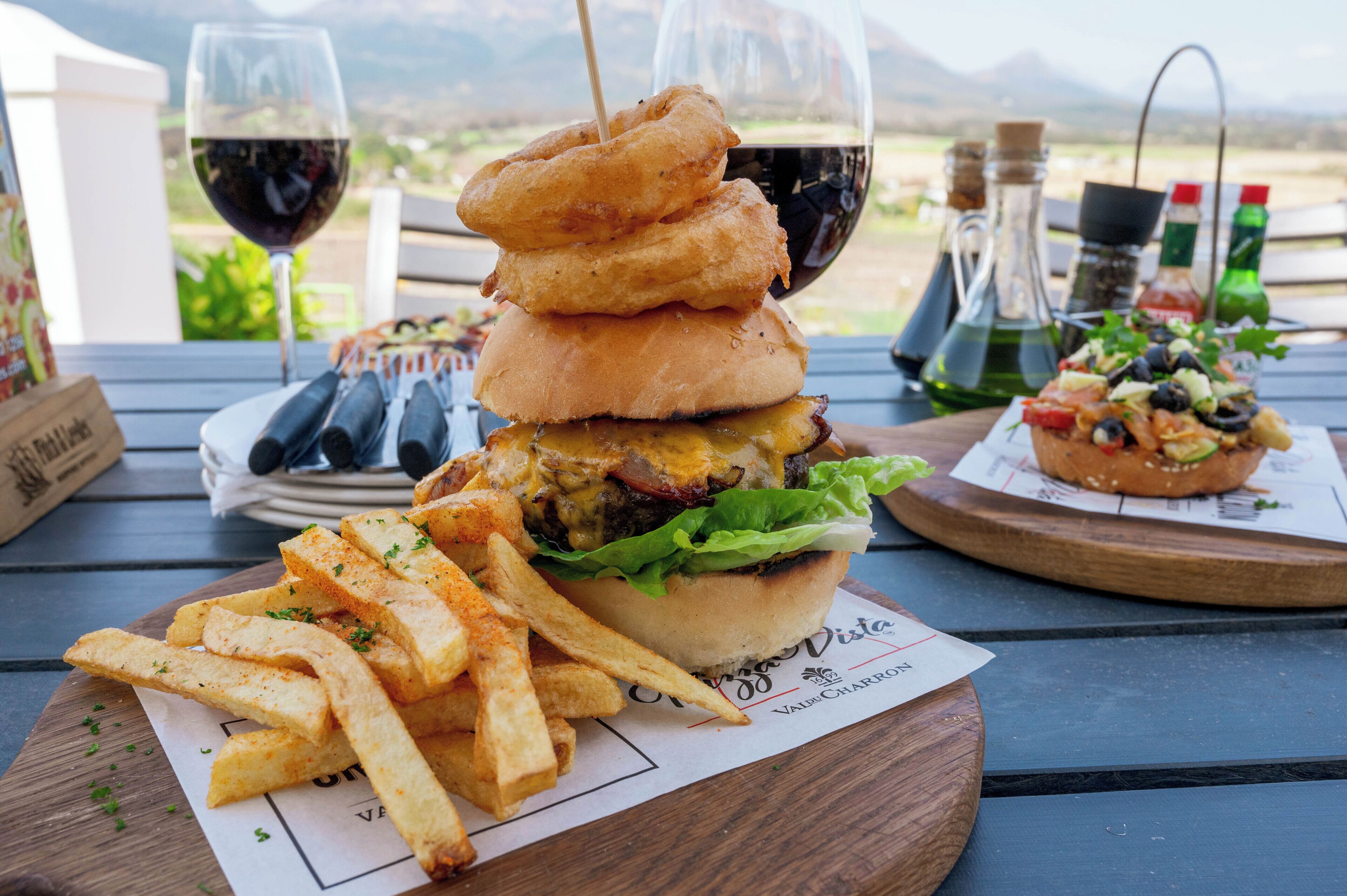 A meal consisting of a burger, fries, and onion rings, served on a wooden table with a view of mountains in the background.