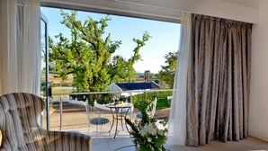 A room with a large window, a striped armchair, a glass coffee table with a vase, and a view of a balcony with outdoor furniture. - Val Du Charron Wine & Leisure Estate (Wellington)