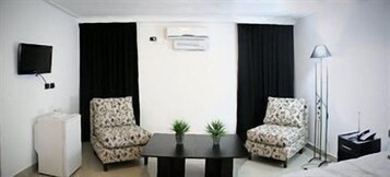 A hotel room with two patterned armchairs, a small table, a TV, an air conditioner, and a floor lamp.
