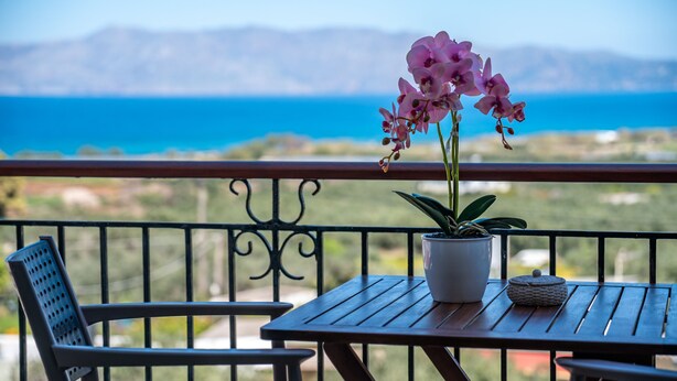 A balcony with a table and chairs, a potted orchid, and a view of the sea and mountains. - Kaliviani Traditional Hotel (Kissamos)