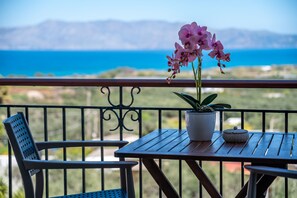 A balcony with a table and chairs, a potted orchid, and a view of the sea and mountains. - Kaliviani Traditional Hotel (Kissamos)