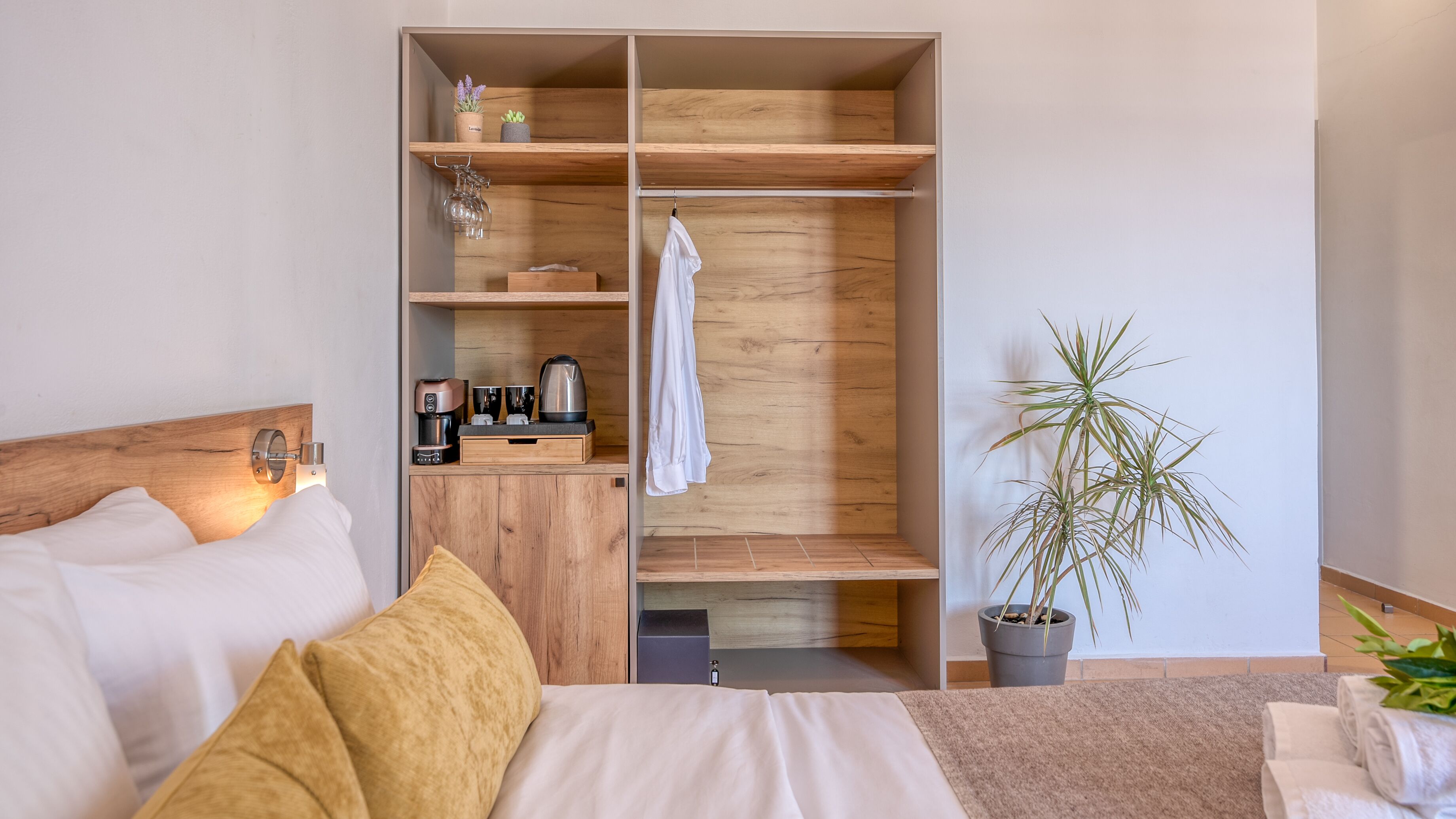 A modern bedroom with a wooden wardrobe, a bed with white linens, a coffee machine, and a potted plant.