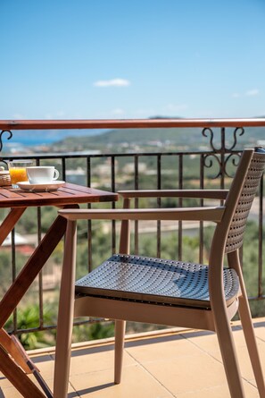 Une table en bois avec une chaise, un verre de jus d’orange et une tasse blanche sur un balcon doté d’une rambarde en métal.