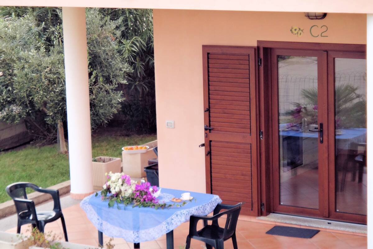 Un patio con una mesa para dos, una puerta con un número y un jardín al fondo.