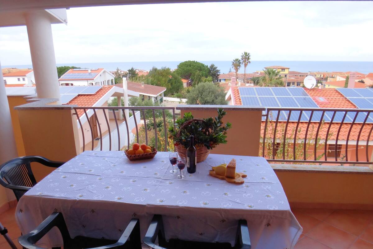 Un balcón con una mesa dispuesta para dos personas, que incluye un mantel, una cesta de naranjas, una botella de vino y una maceta.