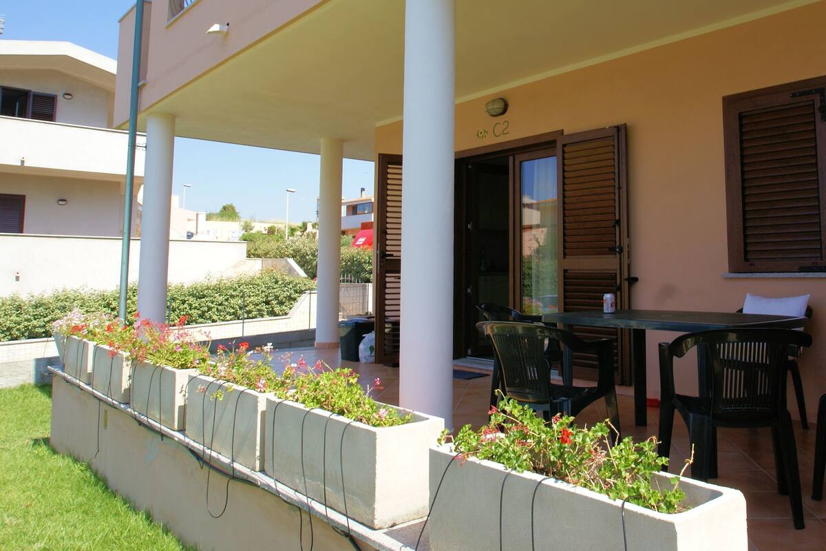 Un espacio al aire libre techado con mesa y sillas, una jardinera con flores y la entrada de un edificio con un letrero.