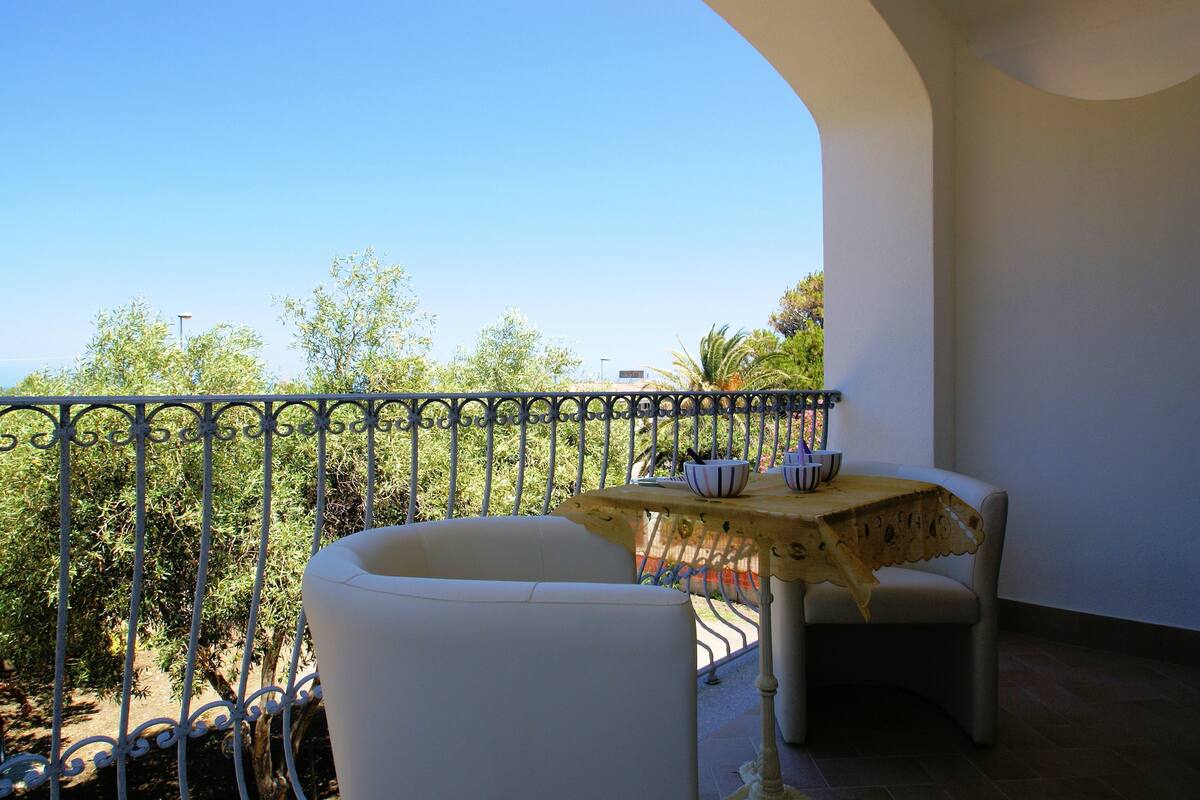 Un balcón con una mesa y sillas, con vista a un jardín con árboles y cielo despejado.
